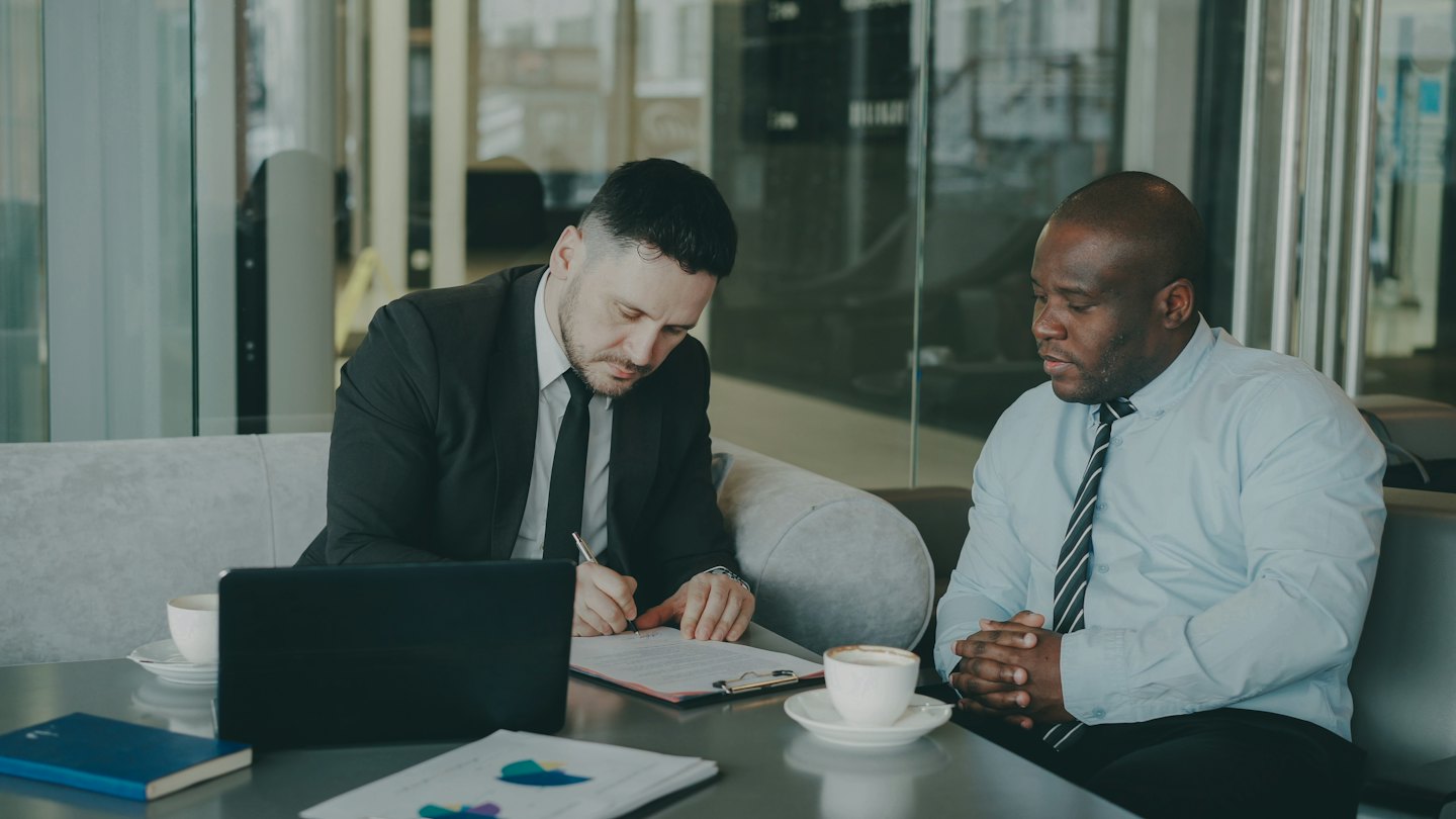 Man in suit signs document while colleague watches.